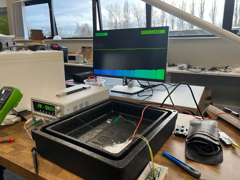 Monitor showing 51C and 800MHz in the background, the Pico 2 in dry ice in the foreground, now connected to a bench supply on the left  The bench supply reads 28V and 600mA